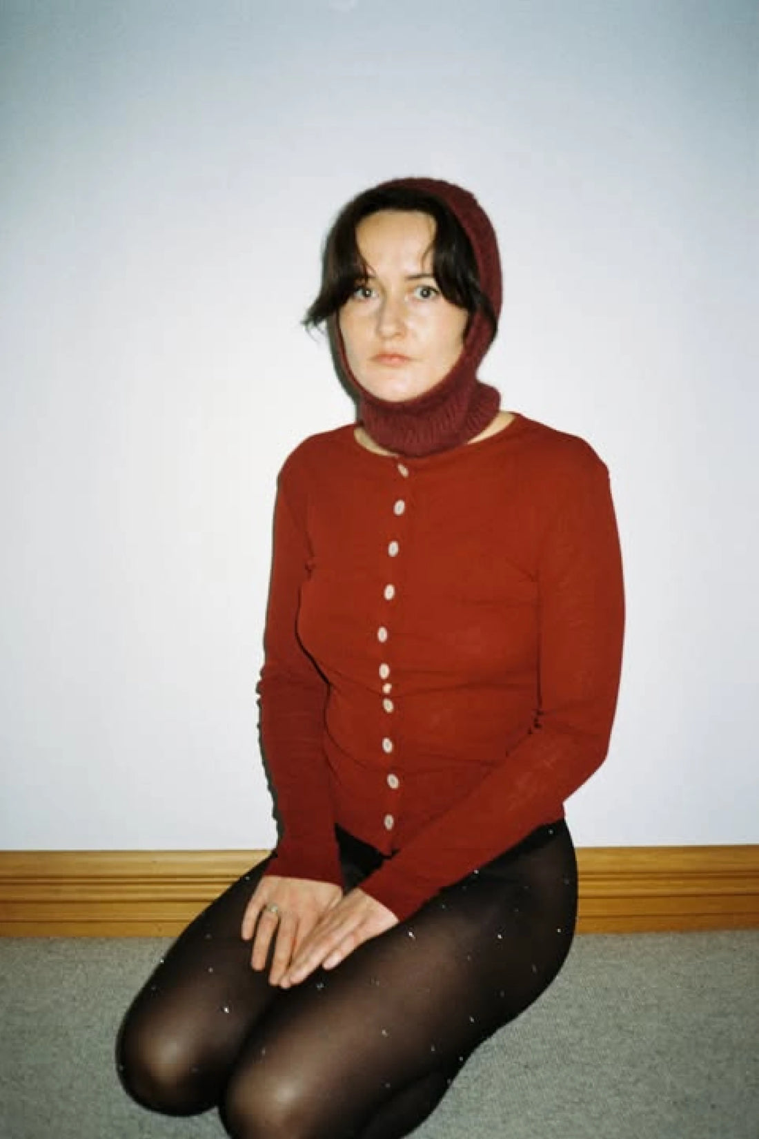 Woman wearing Bonnie Wroe Balaclava - Burgundy and a red cardigan, posed on the floor.