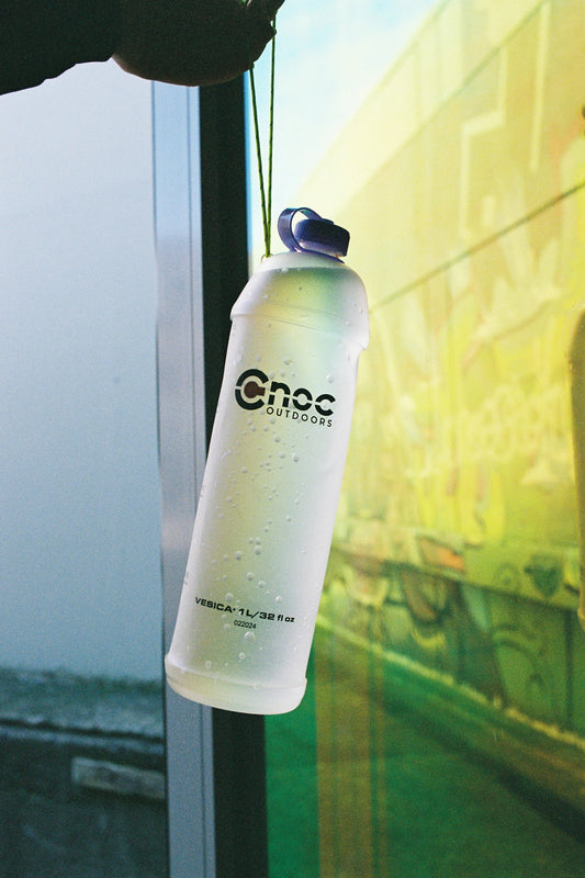 CNOC Vesica 1L Collapsible Bottle 28mm Thread hanging near a colorful wall, showcasing its sustainable design.