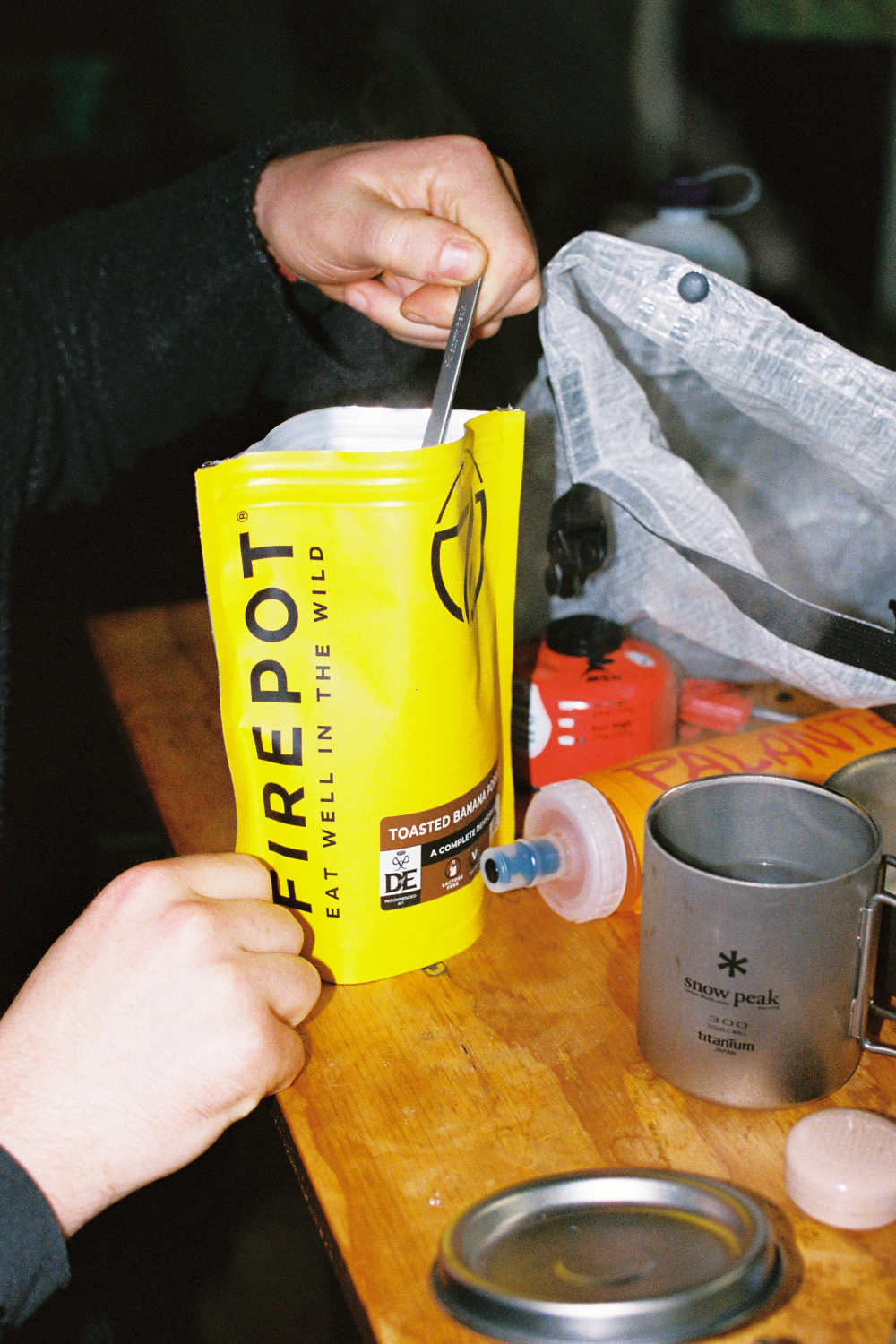 Person preparing Firepot Toasted Banana Porridge from a yellow pouch in an outdoor setting.
