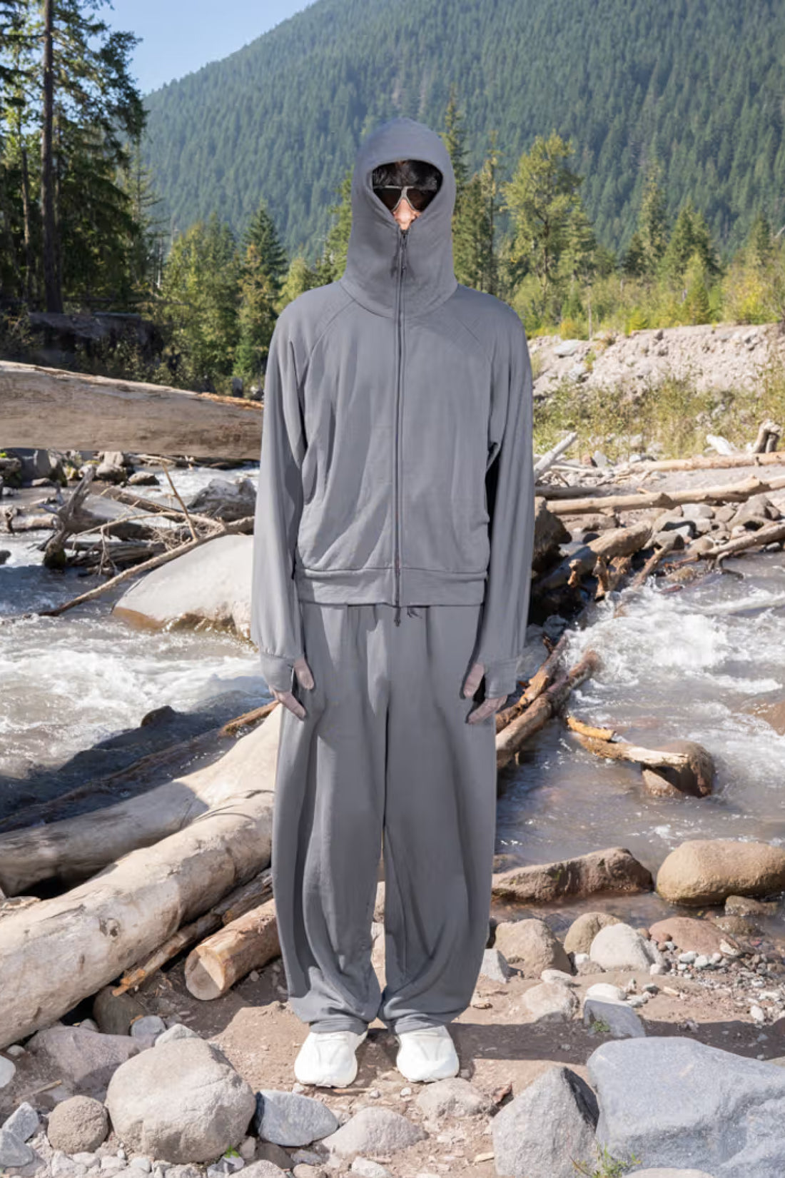 Person wearing Gnuhr Power Pant in Asphalt with a matching hooded jacket, standing by a river and mountains.