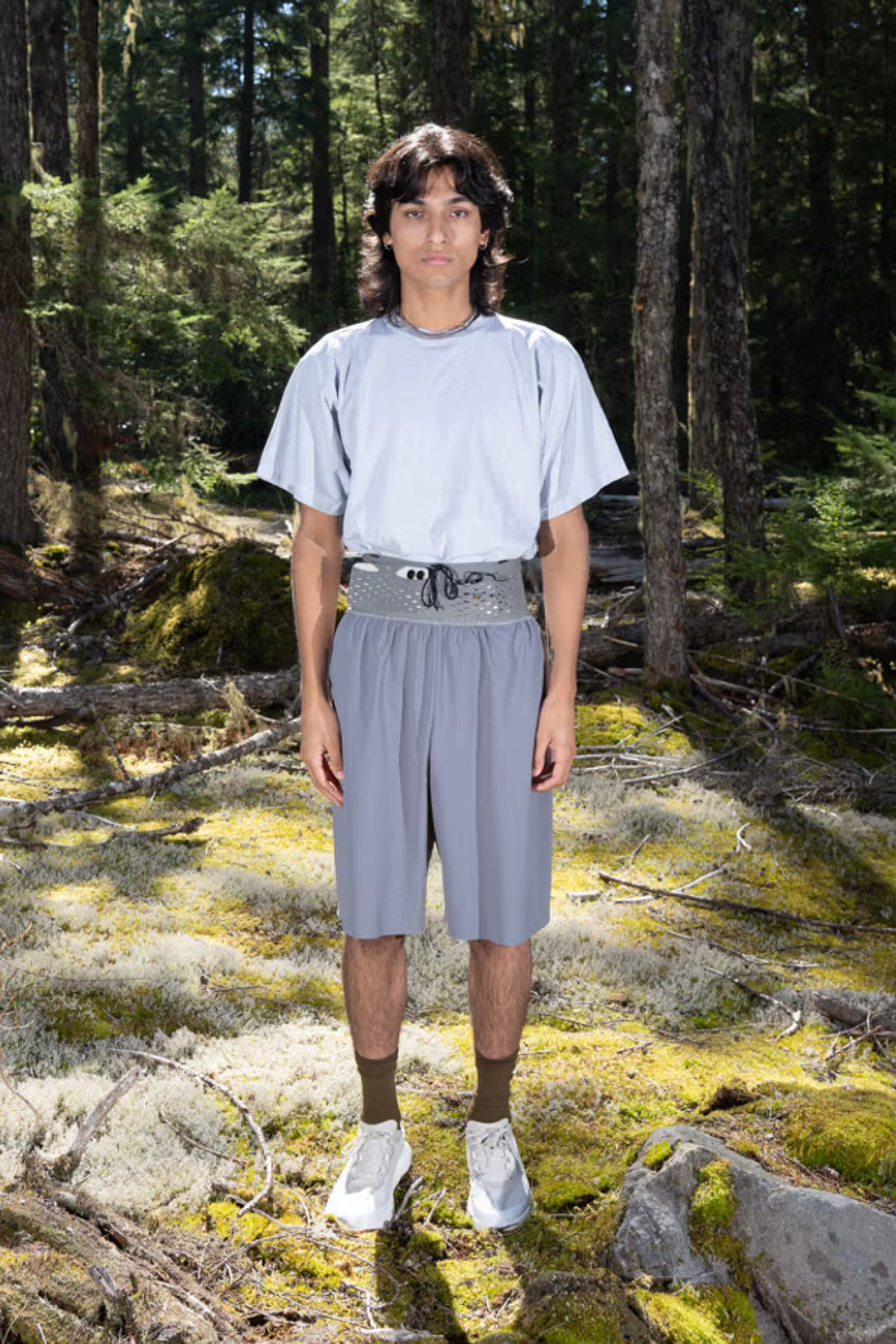 Model wearing Gnuhr Warp Short Long in charcoal color, showcased in a forest setting.