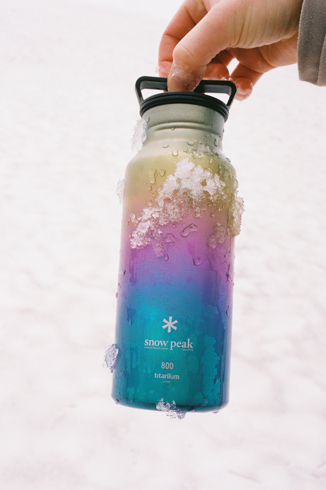 Hand holding the Snow Peak Titanium Aurora Bottle 800 - Rainbow with condensation in snowy background.