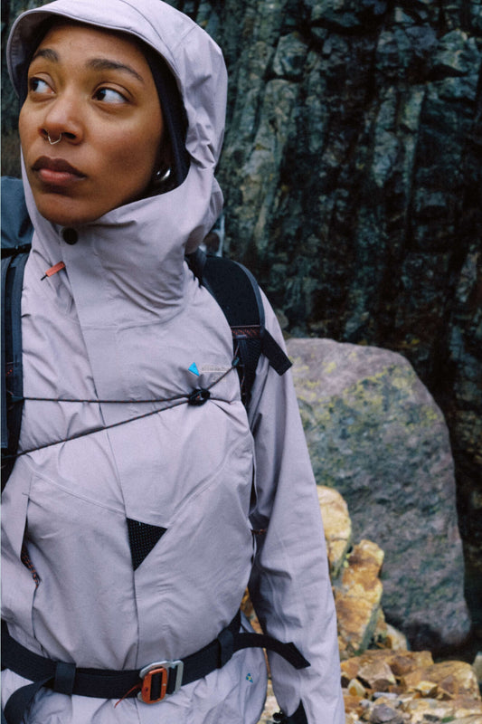 Woman wearing Klattermusen Womens Asynja Jacket in Purple Dove, standing outdoors near rocky terrain.
