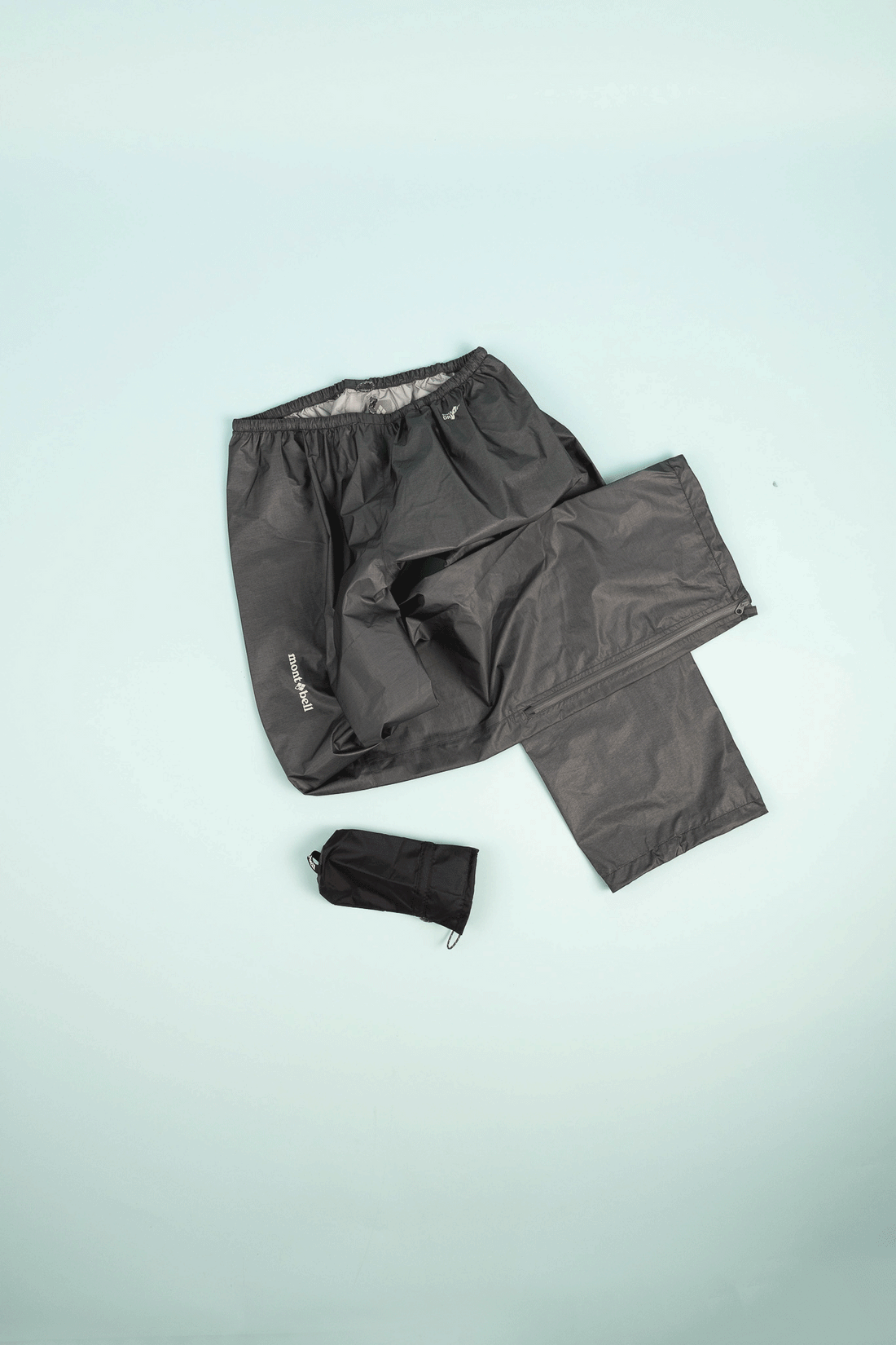 Montbell US Women's Dry-Tec U.L. Rain Pants in dark gray, displayed with storage bag, ideal for ultralight outdoor wear.